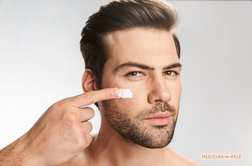 Homem de barba feita aplica creme no rosto com o dedo indicador, olhando diretamente para a câmera.