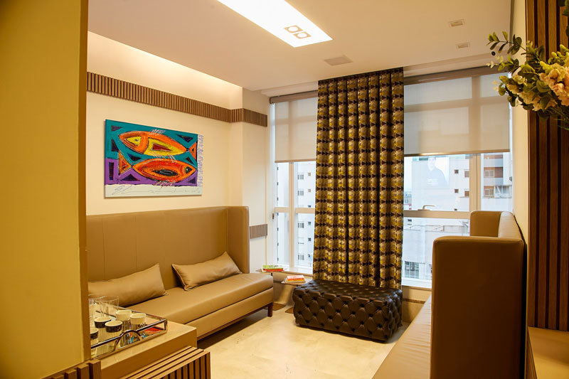 Recepção sofisticada de clínica de dermatologistas em Florianópolis com sofás bege, mesa de centro, cortinas estampadas e quadro colorido na parede.