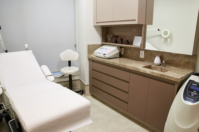 Sala de atendimento dermatológico com maca branca, bancada em mármore, equipamentos e armários.