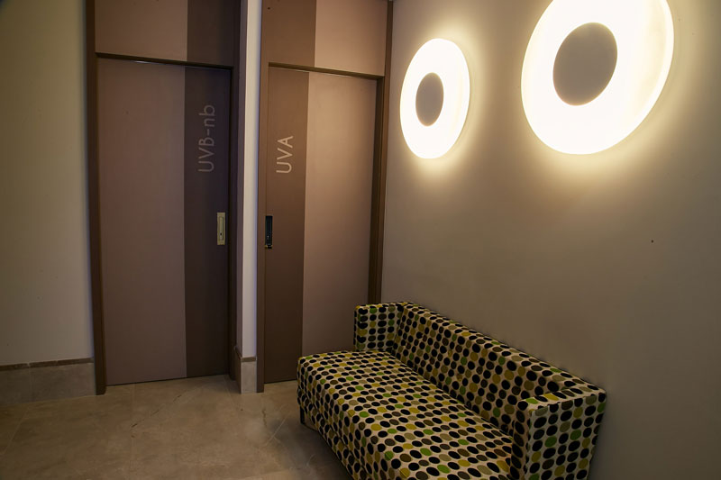 Corredor com sofá estampado em tons de verde, preto e bege, duas portas de consultórios e luminárias circulares na parede de clínica de dermatologistas em Florianópolis.