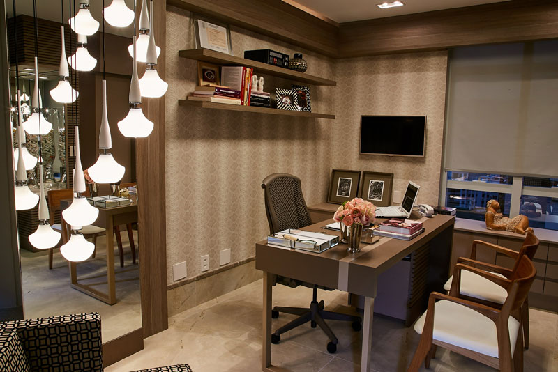 Consultório sofisticado com mesa de atendimento, poltronas, luminárias pendentes, prateleiras com livros e porta-retratos, além de uma janela com persiana.