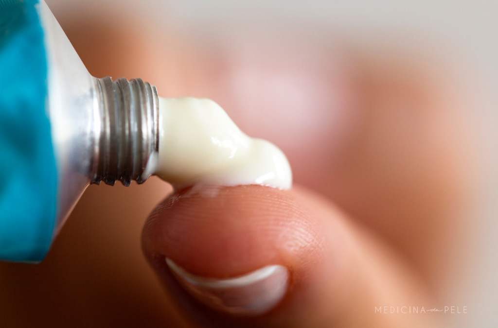 Close de pomada branca sendo aplicada na ponta de um dedo, com o tubo metálico aberto ao lado.