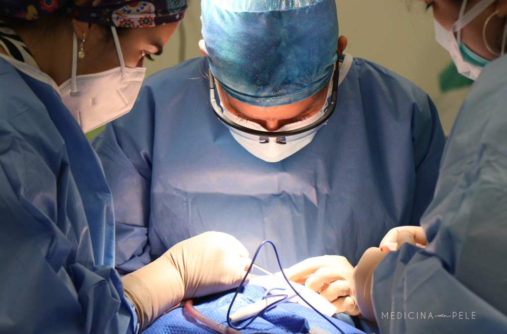 Três profissionais com roupas cirúrgicas e máscaras realizam procedimento médico em centro cirúrgico, concentrados sobre o campo operatório iluminado.