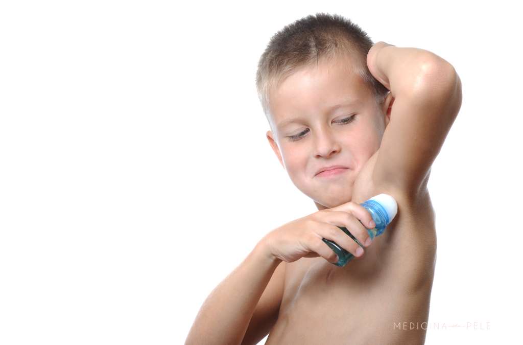 Menino sem camisa aplica desodorante roll-on na axila com expressão concentrada e divertida.