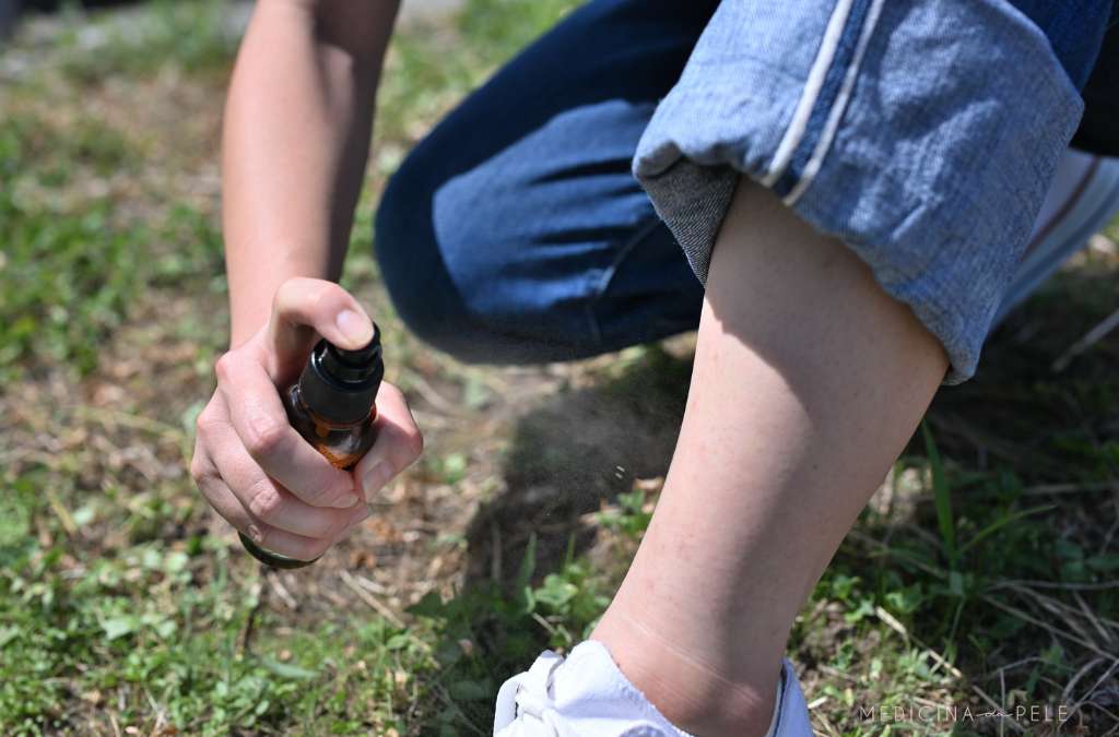 Pessoa aplica spray repelente na perna, ajoelhada sobre grama, com calça jeans dobrada.