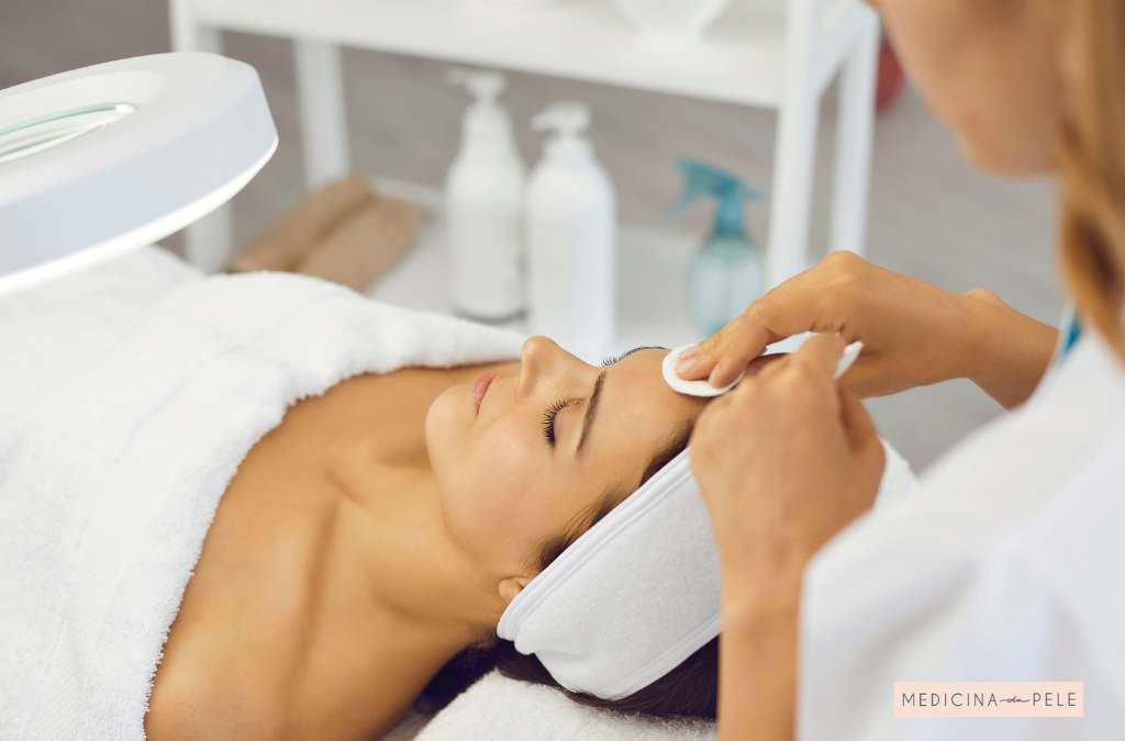 Mulher deitada com toalha branca recebe limpeza facial; profissional usa algodão na testa dela.
