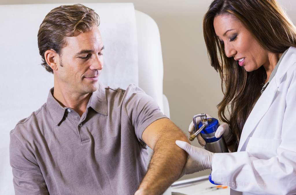 Profissional aplica crioterapia no braço de homem sorridente, usando equipamento com jato de nitrogênio.