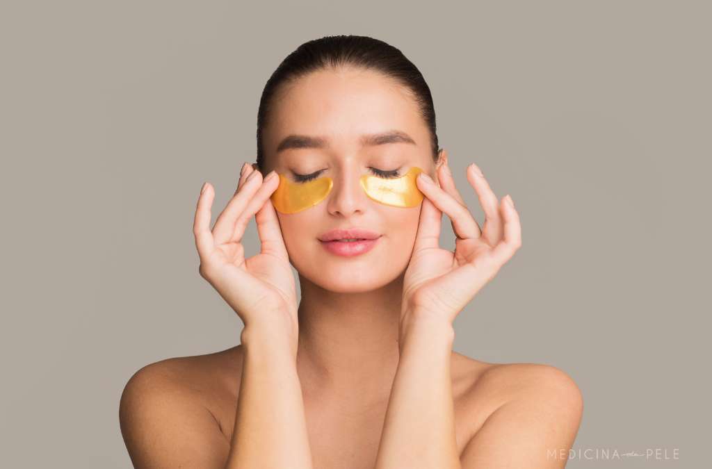 Mulher de olhos fechados sorri enquanto ajusta par de patches dourados sob os olhos em fundo neutro.