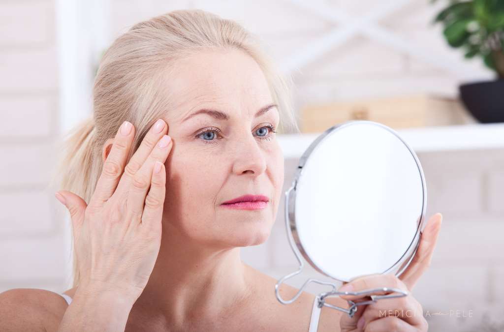 Mulher madura se observa no espelho enquanto toca suavemente a pele ao redor dos olhos com expressão atenta.