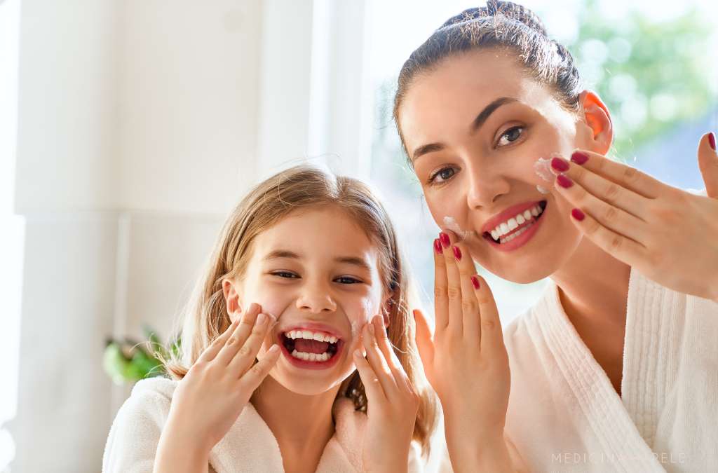Mulher e menina sorridentes aplicam creme facial, usando roupões claros em banheiro iluminado.