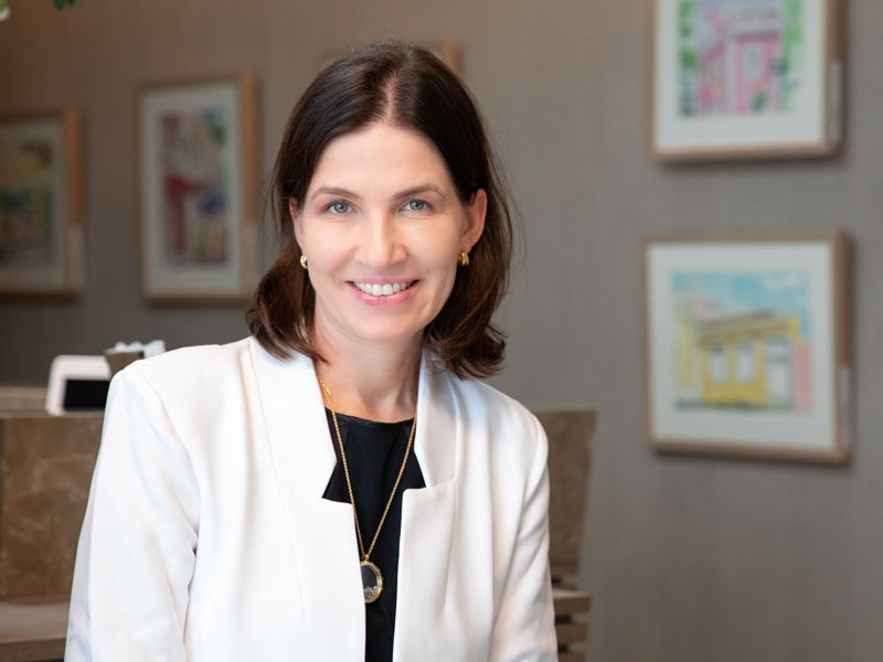 foto de Doutora Tatiana Biasi, dermatologista em Florianópolis: Mulher de cabelos castanhos lisos e curtos sorri levemente; ela veste blazer branco sobre blusa preta e usa colares dourados.