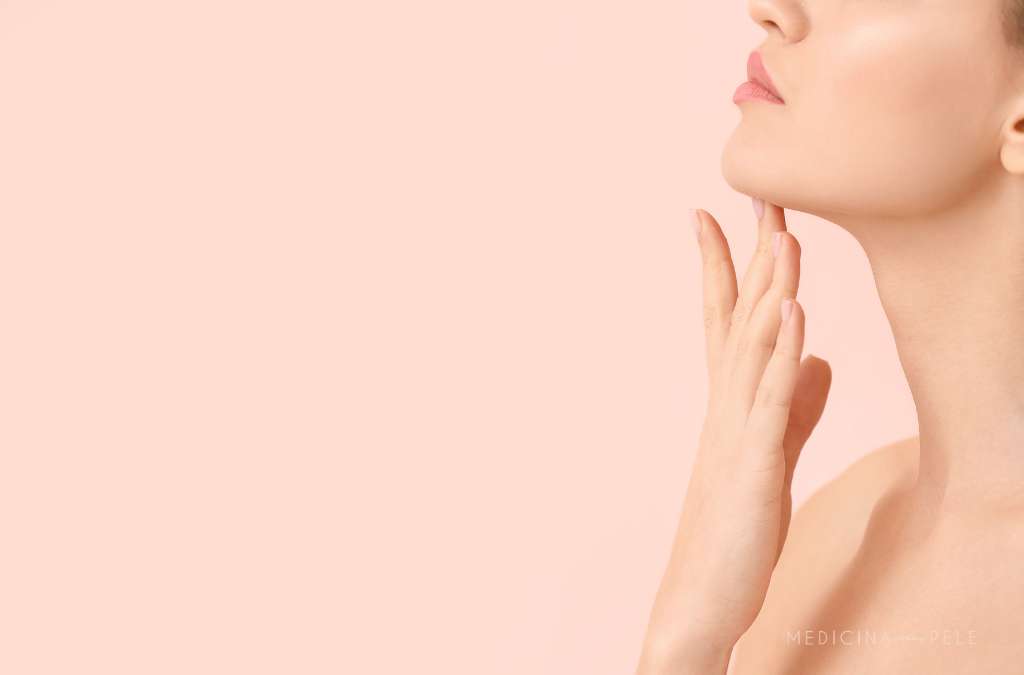 Fotografia mostra uma mulher com pele lisa tocando suavemente a região inferior do rosto e do pescoço, sugerindo cuidados estéticos com foco em definição do contorno facial, como o uso de enzimas lipolíticas ou esvaziador injetável.