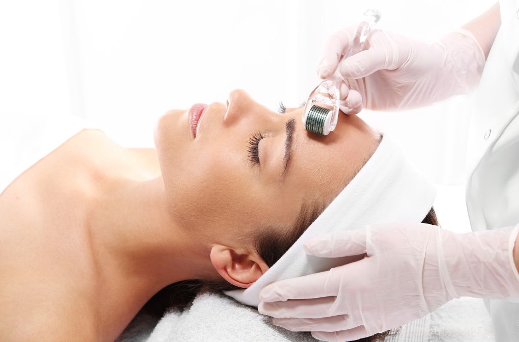 Mulher deitada com faixa branca na cabeça recebe tratamento facial com dermaroller aplicado na testa por profissional usando luvas rosas.