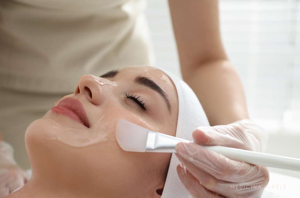 Fotografia mostra uma mulher deitada com faixa na cabeça enquanto profissional aplica substância transparente no rosto com espátula, ilustrando procedimento de peeling químico.