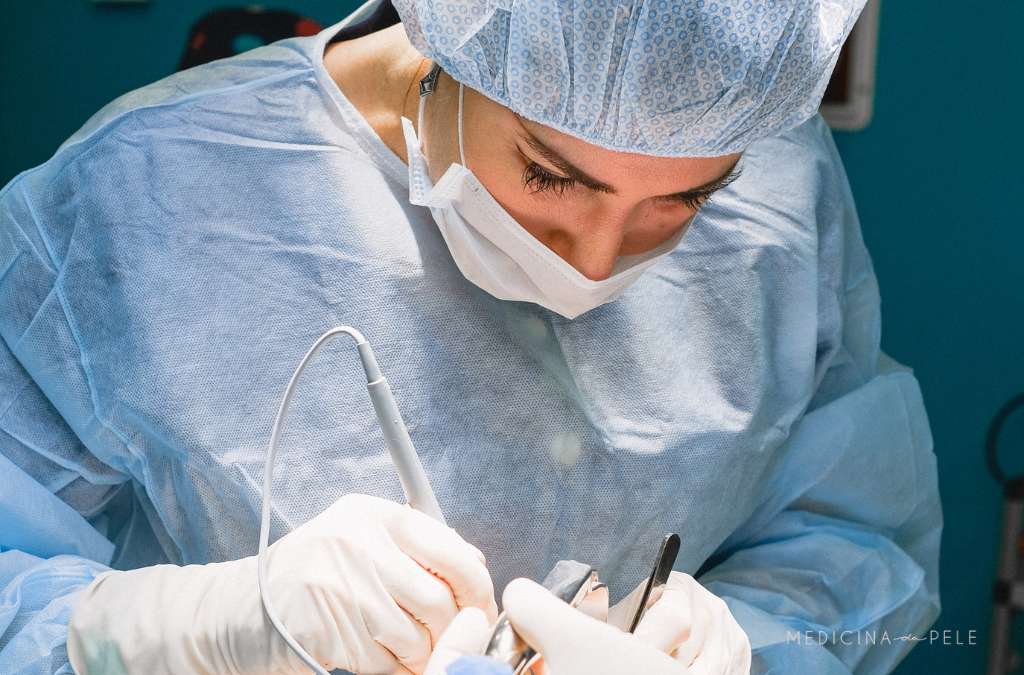 Fotografia mostra cirurgiã com roupas e máscara esterilizadas realizando procedimento minucioso, indicando possível retirada de tumor cutâneo ou cirurgia dermatológica.