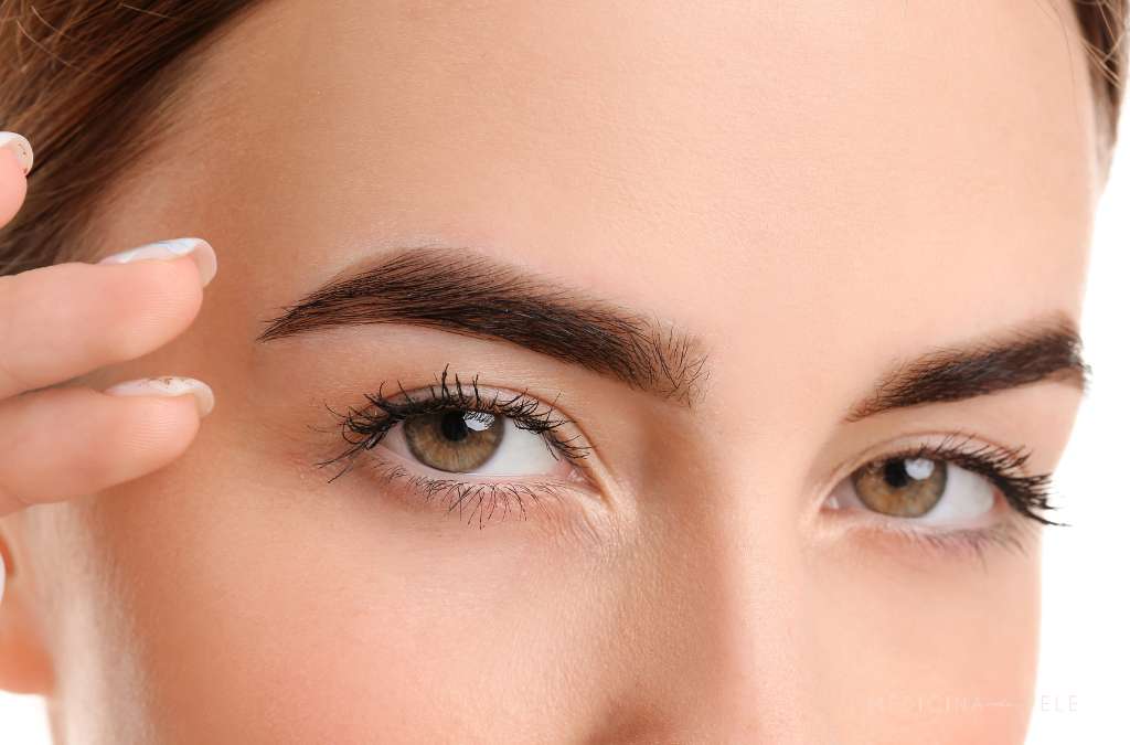 Close do rosto de uma mulher destacando sobrancelhas espessas e bem definidas, cílios alongados e pele uniforme, sugerindo cuidados estéticos com a região dos olhos.