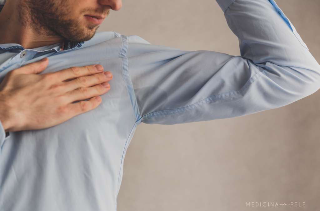 Fotografia mostra um homem vestindo camisa azul clara observando a própria axila, possivelmente preocupado com suor excessivo ou odor.