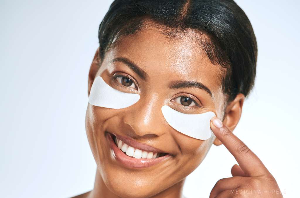 Mulher sorridente com adesivos brancos na região abaixo dos olhos, apontando para um deles; imagem com fundo claro.