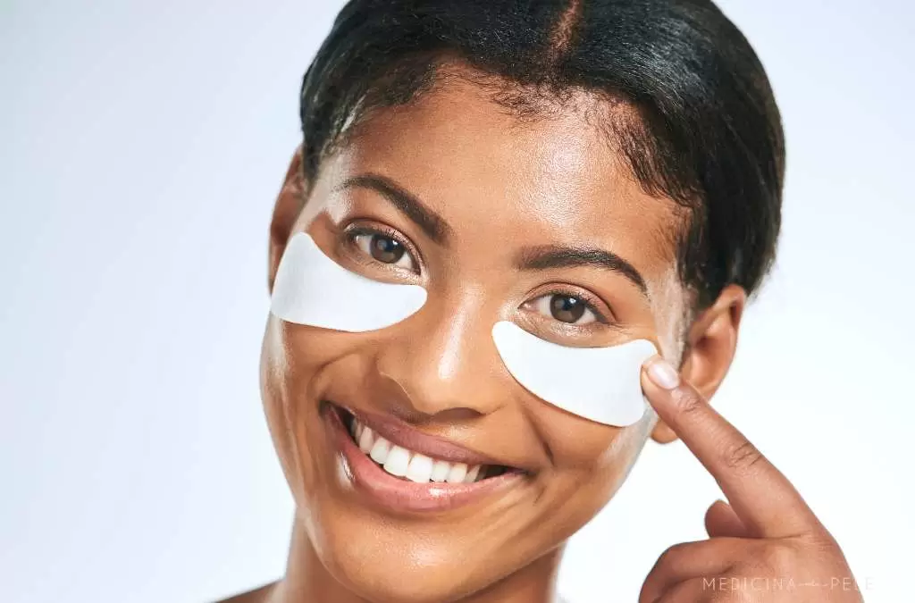 Mulher sorridente com adesivos brancos na região abaixo dos olhos, apontando para um deles; imagem com fundo claro.