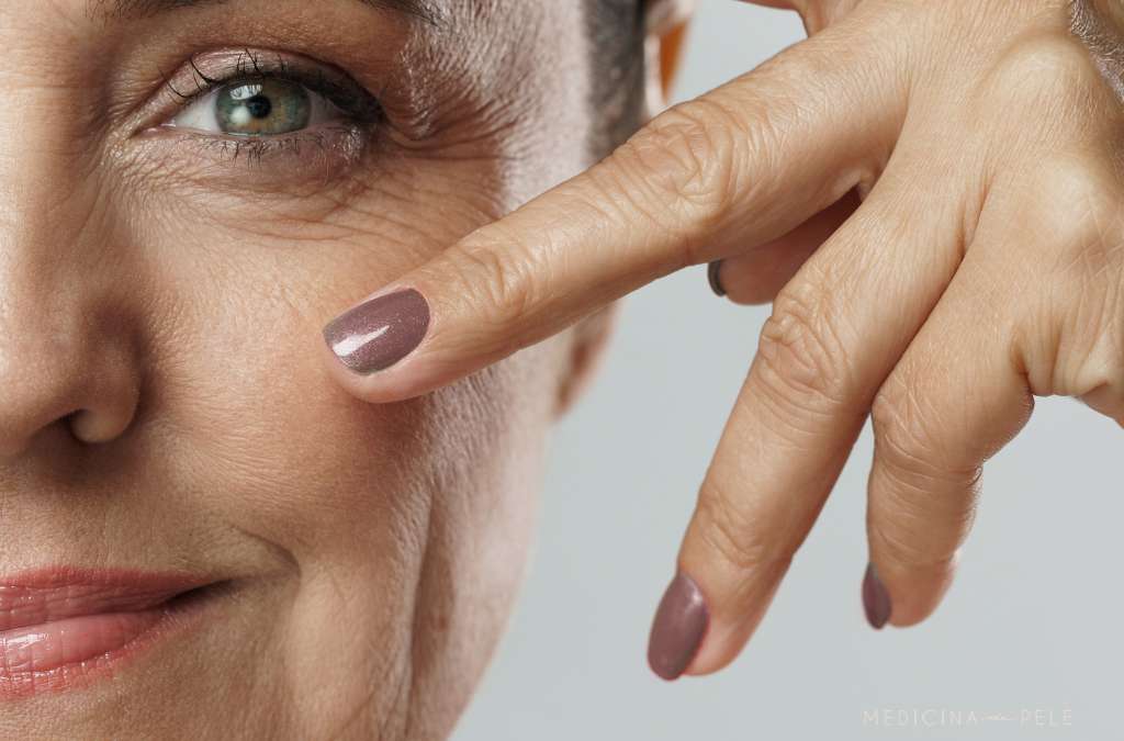 Close-up de mulher madura com expressão serena, apontando com dois dedos para a região abaixo dos olhos, destacando rugas e linhas de expressão.