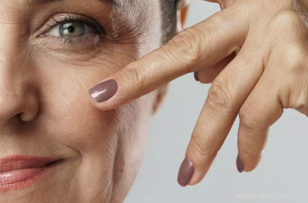 Close-up de mulher madura com expressão serena, apontando com dois dedos para a região abaixo dos olhos, destacando rugas e linhas de expressão.