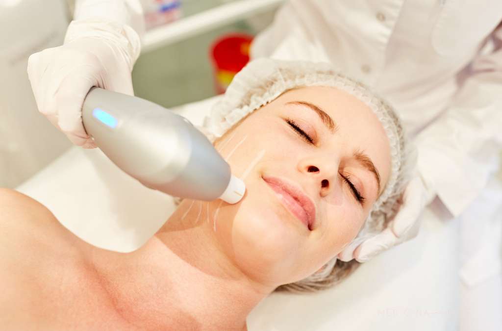Mulher deitada recebe tratamento facial com Ultrassom microfocado; profissional com luvas aplica aparelho na bochecha.