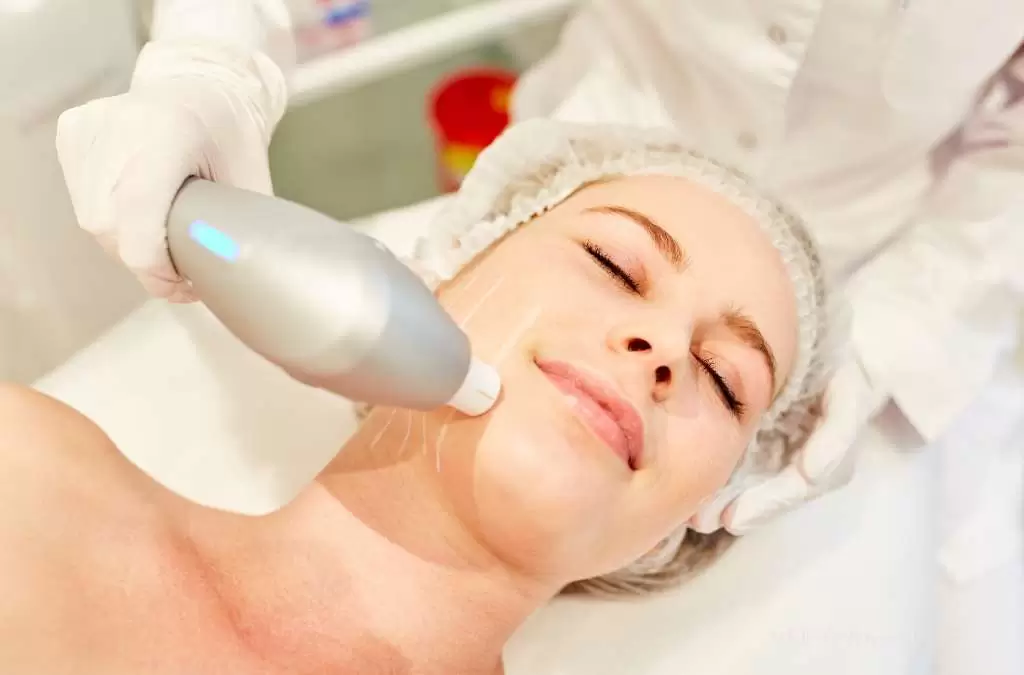 Mulher deitada recebe tratamento facial com Ultrassom microfocado; profissional com luvas aplica aparelho na bochecha.