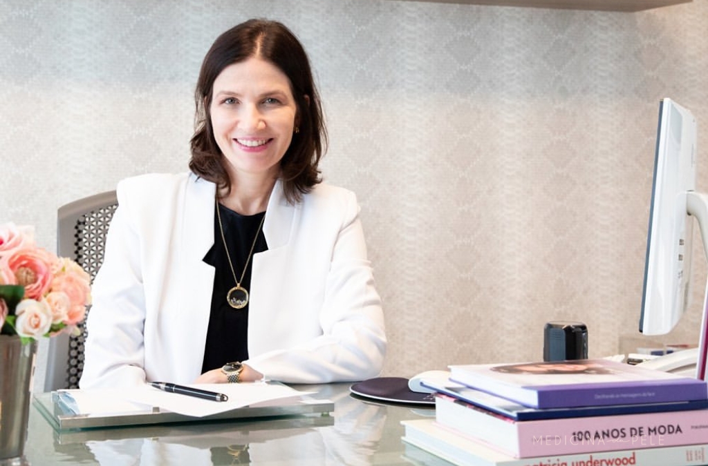 Mulher sorridente de jaleco branco senta-se à mesa com livros e flores, em ambiente de consultório elegante.