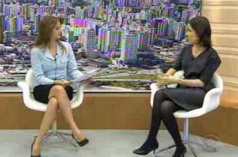 Duas mulheres sentadas em cadeiras brancas conversam em estúdio de TV, com imagem aérea de cidade ao fundo.