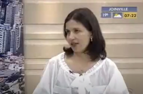 Mulher de blusa branca com laço participa de entrevista em estúdio de TV, com informações de clima e horário na tela.