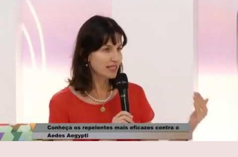 Mulher de blusa vermelha fala ao microfone em estúdio de TV, com legenda sobre repelentes contra o mosquito Aedes aegypti.