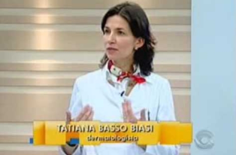 Mulher de jaleco branco e lenço estampado no pescoço fala durante entrevista em programa de TV, com legenda identificando-a como dermatologista.