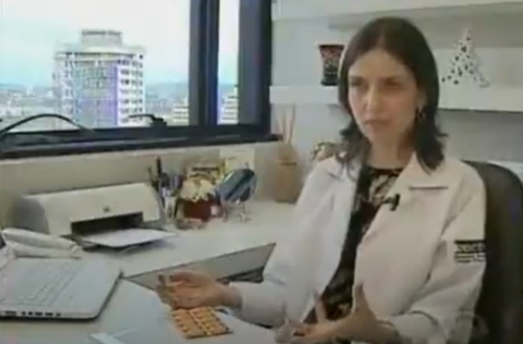 Mulher de jaleco branco sentada em consultório segura cartela de comprimidos enquanto fala, com janela mostrando vista urbana ao fundo.