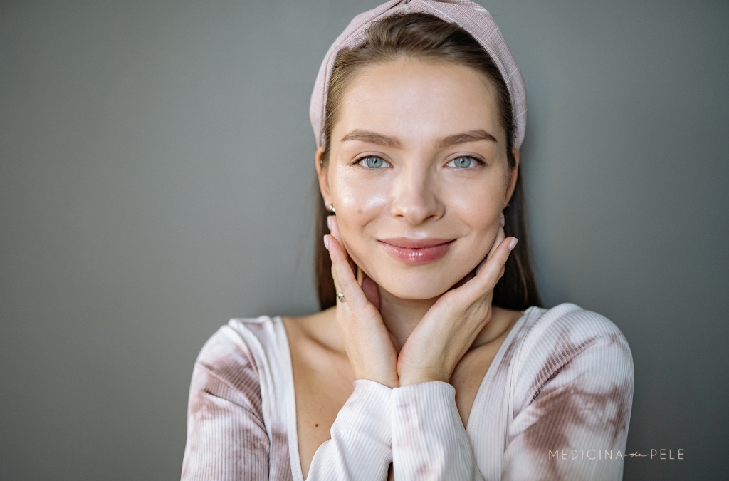 Mulher de pele clara e olhos claros sorri suavemente, apoiando o rosto nas mãos, usando faixa de cabelo rosa.