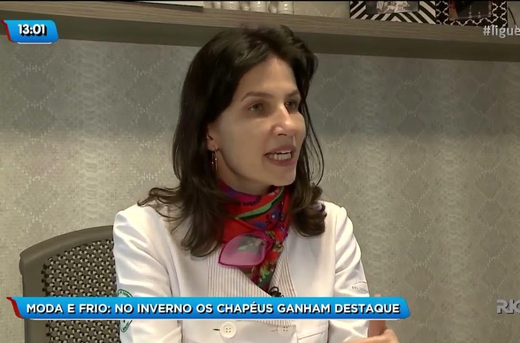 Fotografia de estúdio mostrando a Dra. Tatiana Biasi, dermatologista, com expressão atenta e gesticulando enquanto fala. Ela veste blazer branco e lenço estampado em vermelho e verde. Na parte inferior da imagem, há uma faixa com o texto: “MODA E FRIO: NO INVERNO OS CHAPÉUS GANHAM DESTAQUE”. Ao fundo, uma estante com objetos decorativos.