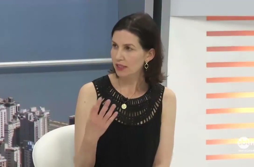 Fotografia de estúdio televisivo mostrando uma mulher de cabelos castanhos escuros e lisos, usando vestido preto sem mangas e brincos dourados. Ela está sentada, gesticulando com a mão direita enquanto fala. Ao fundo, há uma imagem urbana com prédios e mar ao lado esquerdo, e painéis com listras laranja à direita.