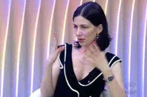 Mulher de vestido preto com detalhes brancos gesticula enquanto fala durante participação em programa de TV.