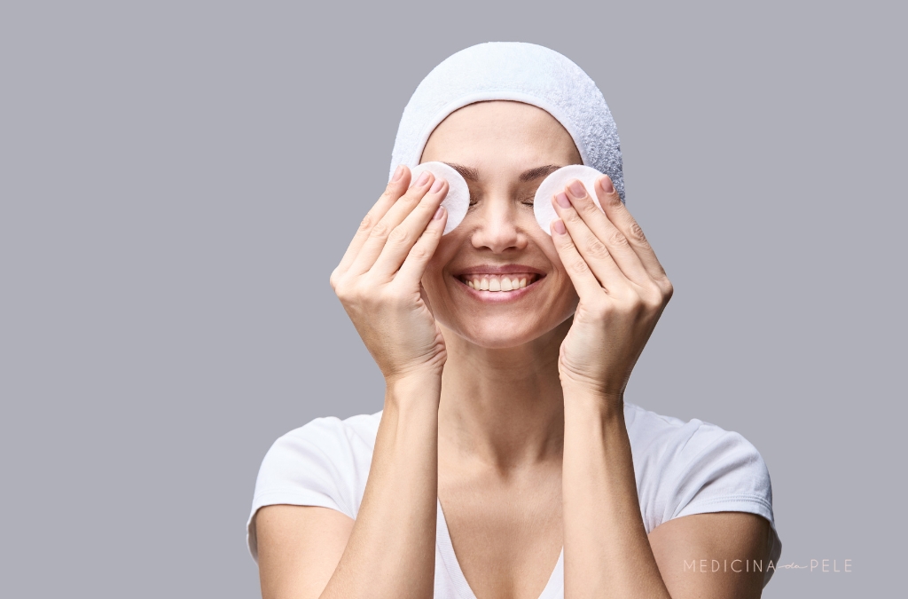 Mulher sorridente com toalha na cabeça segura discos de algodão sobre os olhos, em fundo cinza neutro.