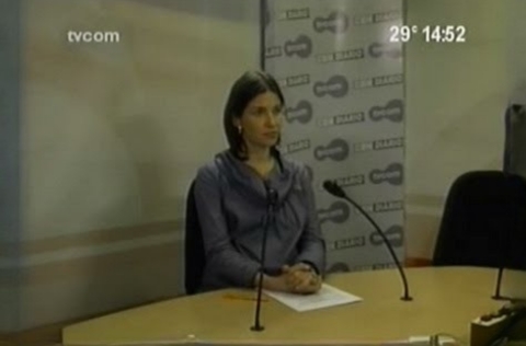 Mulher sentada em estúdio de rádio durante entrevista, com microfones posicionados à sua frente e logotipos ao fundo.