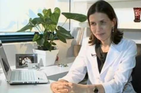 Mulher de jaleco branco sentada à mesa em consultório, com laptop e vaso de planta ao fundo, olhando atentamente para a frente.