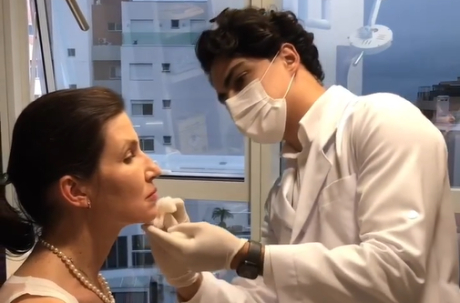Profissional de jaleco e máscara realiza procedimento estético no queixo de paciente.