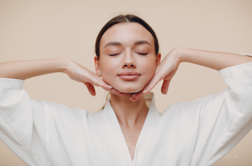 Mulher de olhos fechados e expressão relaxada apoia o queixo nas mãos, vestindo robe branco.