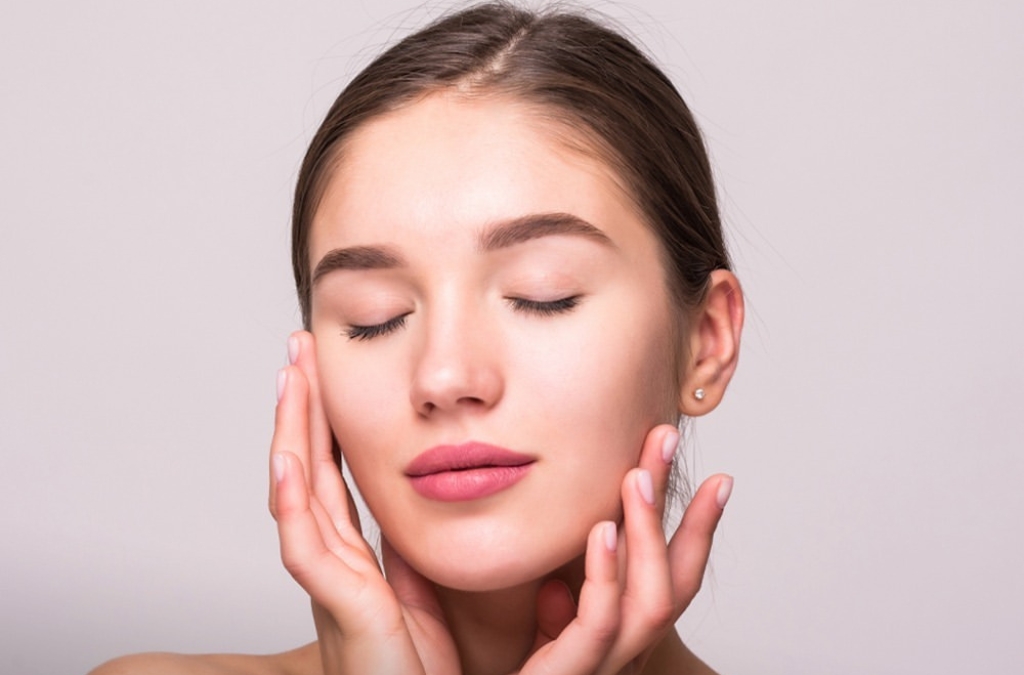 Mulher jovem de olhos fechados e expressão serena, tocando suavemente o próprio rosto com as duas mãos, sobre fundo claro, acompanhada da legenda “Procedimento Profhilo” e a marca “Medicina da Pele” na parte superior.