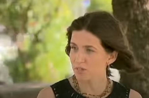 Mulher de vestido preto fala em ambiente externo, com expressão séria e colar dourado, tendo árvores desfocadas ao fundo.