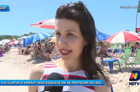 Mulher de vestido listrado concede entrevista na praia, com guarda-sóis coloridos ao fundo e legenda sobre proteção solar.