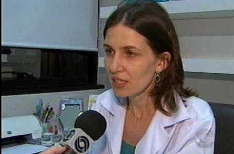 Mulher de jaleco branco concede entrevista a repórter que segura microfone com logotipo de emissora de TV.