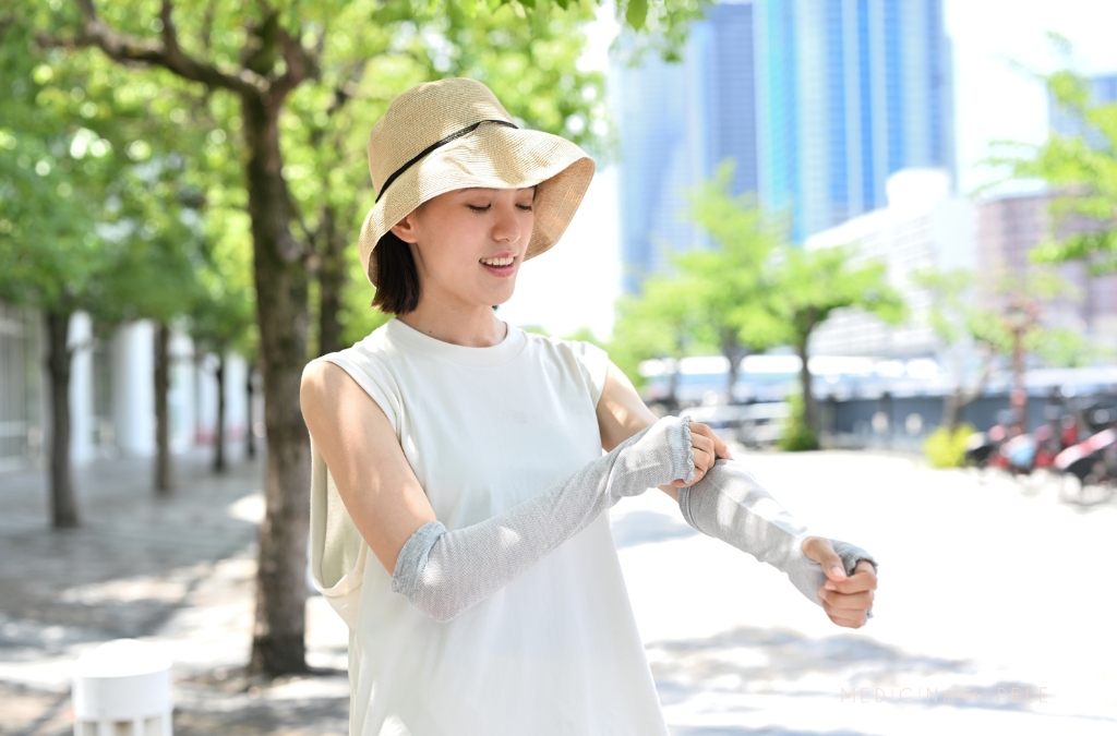 Mulher sorridente ao ar livre usa chapéu de abas largas e veste manguitos de proteção solar nos braços.