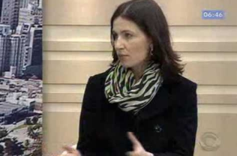 Mulher com casaco preto e lenço estampado participa de entrevista em estúdio de TV, gesticulando enquanto fala.