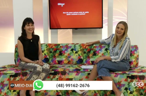 Duas mulheres sentadas em sofá colorido conversam sorrindo em estúdio de TV, com telefone de contato exibido na tela.