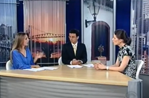 Dois apresentadores e uma entrevistada conversam em estúdio de TV, sentados à mesa com papéis e canetas.